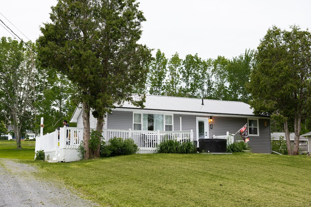 The grey house with its white front porch and hot tub, on a wide lawn between tall trees